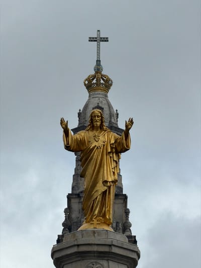 A low-angle, eye-level shot captures a golden statue of Jesus Christ against an overcast sky and the upper portion of a grey stone church tower. The statue depicts Jesus standing, facing forward, with long hair, a beard, and a mustache. Both arms are raised and slightly bent at the elbows, with palms open and fingers slightly splayed upwards. A detailed Sacred Heart emblem, featuring flames, a cross, and thorns, is visible on the statue's chest. The figure is clad in a flowing, draped robe. The statue rests on a multi-tiered, grey stone pedestal. Directly behind and above the statue, the ornate grey stone tower of the Basilica of Our Lady of the Rosary of Fátima rises, topped by a large golden crown, and further above, a simple metal cross. The background consists of a uniformly cloudy, light grey sky. The scene is located within the city of Fátima, Portugal, specifically at the Sanctuary of Fátima.