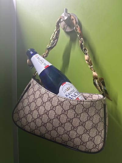 A purse with a patterned design is hanging from a metal hook on a green surface. A beer bottle is inside the purse. The visible text on the bottle reads "KRONE BLANCHE BIERE" and "WITH A HINT OF CITRUS". The location is not discernable from the image.