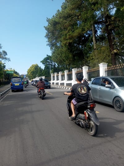 The image depicts a street scene during the daytime. The sky is clear blue. Several vehicles, including cars and motorcycles, are moving along the road. A young person rides a motorcycle, wearing a black t-shirt with a circular design on the back and purple shorts. A silver car is to their right. Further ahead, there's a blue van. A fence borders the street on the right, with trees behind it. A street sign indicating "Istana Cipanas" is visible on the left side of the image.
