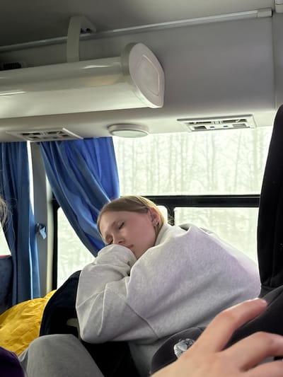 A young woman is sleeping on a bus, with her head resting against the window. She is wearing a gray hooded sweatshirt. A blue curtain is visible behind her head, and the interior of the bus, including an air vent and a white panel, are also visible. The location of the image is unknown and cannot be ascertained based solely on visual information. There is no visual information that suggests the city of Ivanovka, Russia.