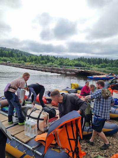 A group of people are on a wooden raft, preparing for an excursion on a wide river. The river is bordered by a dense forest of green trees and the sky is overcast with clouds. Several inflatable rafts are moored to the wooden raft. People are seen handling bags and gear. One woman is wearing a light pink t-shirt and dark pants, with a purple flotation device around her waist. Another person, wearing a black and white patterned top, is leaning over, interacting with the gear. A man in a grey hoodie is looking down at the supplies, and another man in a blue plaid shirt is walking away from the camera. Various bags, water bottles, and life vests are visible on the raft. In the background, a rustic wooden bridge or walkway extends across the river, supported by pontoons. The ground on the riverbank is covered in small stones and gravel.