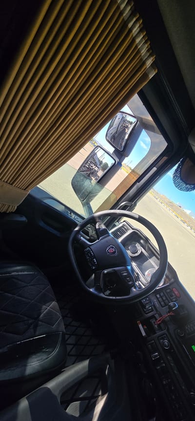 The image shows the interior of a truck cabin, specifically the driver's side, looking out the windshield and side window. A brown, pleated curtain is drawn across the top of the windshield. The steering wheel, with the Scania logo, is in the foreground, along with a portion of the dashboard and driver's seat. The dashboard features various buttons, switches, and a display screen. The driver's seat is black and quilted.

Through the windshield and side mirror, we can see the exterior environment. The side mirror reflects another truck, and further out, the landscape appears to be an open, paved area, possibly a parking lot or an industrial zone, with yellow markings and some distant structures under a bright sky.

The image is taken from the perspective of someone inside the truck, looking forward and slightly to the side. The lighting suggests it is daytime, with sunlight illuminating the scene.

Based on image metadata, the location is the **Service Area El Olivo, Kilometer 325 of the A-2, in the municipality of Pedrola, province of Zaragoza, Spain.**