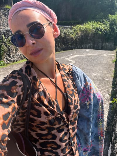 A woman with a shaved head, wearing a pink headband and round sunglasses, poses for a picture outdoors. She is wearing a leopard-print shirt and a tie-dye blue shawl. She is standing on a concrete walkway, with a wall on one side and a cliff face covered in greenery on the other. The location appears to be in or around Benoa, Indonesia, based on the lush, tropical vegetation.