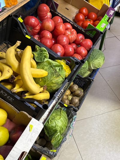 The image depicts an interior view of a produce display within a small grocery store or market in Suhache, Bulgaria. Multiple black plastic crates and cardboard boxes are stacked vertically on a light-colored tiled floor with visible grout lines.
In the upper-left, a black crate contains numerous large, ripe red beefsteak tomatoes. Two yellow price labels are affixed to the crate's edge, one displaying "3.50лв" and another "2.00лв". Adjacent to this, in the upper-right, another crate, partially obscured by a cardboard divider, also holds red tomatoes, some appearing smaller and rounder. A green-branded cardboard piece, possibly reading "SON", is visible among these tomatoes.
Below the tomato crates, a black crate on the left is filled with bunches of ripe yellow bananas. To its right, a crate contains several large green cabbage heads, one of which is partially enclosed in a yellow plastic bag.
Further down, on the bottom left, a cardboard box, partially obscured, displays yellow and red apples. A yellow price label is partially visible on a cardboard divider within this box. Directly to its right, a black crate is filled with unwashed potatoes of various sizes. A yellow price label is attached to the front edge of this potato crate.
In the blurred background, above the produce display, orange-colored liquid bottles are visible on a wooden shelf. No individuals or active interactions are present in the scene; the focus is solely on the static arrangement of fresh produce for retail.