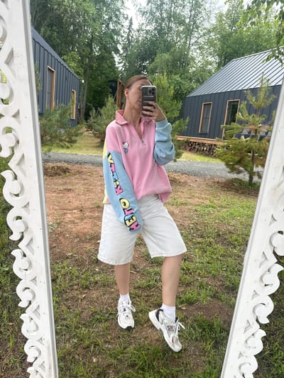 A young woman is reflected in a white, ornate mirror, taking a selfie with her phone. She is wearing a pink and light blue sweater with a Hello Kitty graphic on the sleeve, white denim shorts, and white sneakers. She stands on a grassy area with dirt patches in front of two modern, dark-colored cabins with pitched roofs. The background consists of green trees and foliage, suggesting an outdoor, possibly rural or vacation setting. The mirror is positioned to show her from the waist down, capturing the reflection of the cabins and trees behind her. The scene appears to be in Novaya Vozhoika, Russia, given the context provided.