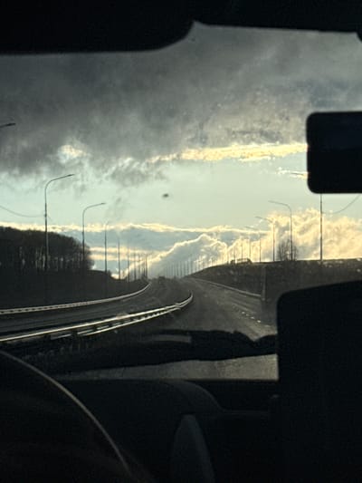 The image captures an outdoor scene from the interior of a moving vehicle, looking out through a window. The view is framed by dark shapes of the vehicle's interior on the left, right, and partially at the bottom.
The central subject is a multi-lane highway or major road that curves gently to the right and extends into the distance. The road surface appears dark, possibly asphalt, with clear white lane markings and solid white lines delineating the shoulder or barrier. Along the left side of the road, a series of streetlights with curved arms are evenly spaced, appearing smaller further down the road. Guardrails or barriers separate the road from the adjacent landscape.
Above the road, the sky is partially cloudy, with distinct, cumulus-like clouds. The lighting suggests either late afternoon or early morning, as parts of the clouds are brightly illuminated by soft, warm light from behind or above, creating a contrast with darker cloud formations. The overall impression is of a clear but partly overcast day.
On the right side of the road, beyond the guardrail, there is a dark stretch of land with what appears to be trees or dense foliage.
No people or vehicles other than the one from which the photo is taken are clearly visible. There is no visible text. The location context provided is Aleksandrovskoe, Russia, indicating a road in that general area.