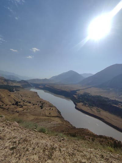 The image depicts a vast, arid landscape under a bright, hazy sun. In the foreground, a rocky, dirt embankment slopes downwards, covered with sparse dry grasses. A wide, muddy river curves through the valley, its banks lined with dry, scrubby vegetation and scattered trees. The valley floor is a patchwork of dry fields, some appearing tilled, others covered in low brush. Beyond the valley, layers of hazy blue mountains recede into the distance, creating a sense of immense depth. The sky is a pale blue, with a few wispy clouds and the sun prominently visible in the upper right corner, casting a strong glare. The scene suggests a remote, undeveloped area, likely in a mountainous region with a dry climate. It is likely located in the Aligalabon area of Tajikistan, given the geographic context. There are no visible people or specific man-made structures besides what appears to be small scattered settlements and possibly roads in the distance.