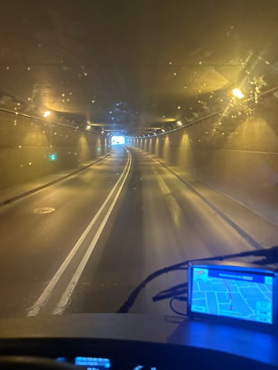 The image shows the interior of a vehicle driving through a tunnel in Memmingen, Germany. The tunnel walls are lit by yellow and white lights. There are green illuminated signs on the left wall. The road surface appears wet, with white lane markings visible. In the foreground, a GPS device is mounted on the dashboard, displaying a map. The windshield has streaks and smudges, indicating it may be wet or dirty. The perspective suggests the photo was taken from the driver's seat.