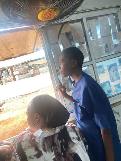 Two people, one wearing a blue shirt and the other wearing a patterned shirt with a black head covering, are looking at a window covered with pictures of faces, likely in a barber shop. A fan is visible in the foreground. The environment outside the window includes corrugated metal roofs and walls, indicative of a low-income area in Okengwe-Obehira, Nigeria. There is bright sunlight outside.
