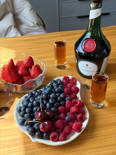 This image depicts a table setting on a wooden surface. The arrangement features a clear glass bowl filled with strawberries, a white plate overflowing with blueberries and raspberries, and two small shot glasses containing an amber liquid. Dominating the right side of the frame is a dark green bottle of Benedictine liqueur, sealed with a red wax emblem. The background suggests an indoor setting with a blurred view of a light-colored puffer jacket on a chair and cabinetry. The lighting indicates a bright, possibly sunlit environment, casting shadows on the table. The location of Борисьево Днп, Russia, is not visually represented in the image.