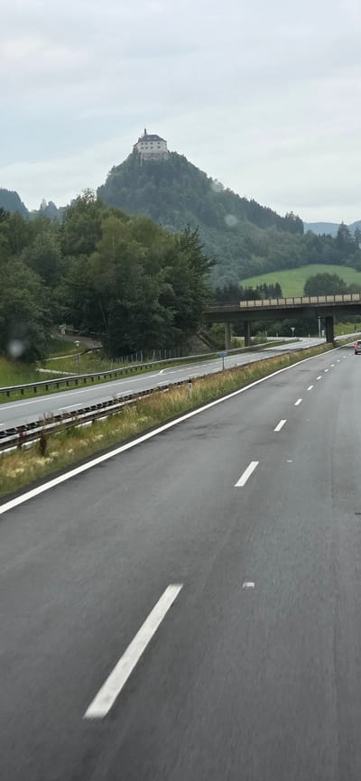 An overcast, grey day frames a view from a highway in Rottenmann, Austria. A large, multi-story building with a prominent tower sits atop a forested hill in the distance, likely a castle or similar historic structure. A multi-lane highway stretches into the distance, with wet asphalt indicating recent rain. White lane markings are clearly visible on the road. A pedestrian overpass crosses the highway in the middle ground, supported by concrete pillars. Green trees and rolling hills are visible on either side of the highway, with some patches of green grass on the hills. A single red car is visible on the highway in the distance, heading away from the viewer. The scene suggests a travel or transit moment, capturing the unique landscape of the Austrian region.