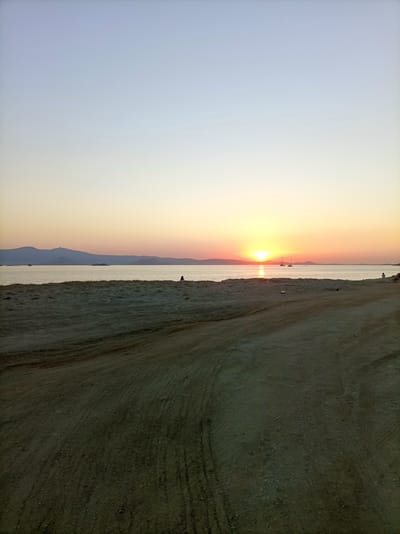 A serene beach scene unfolds at sunset on Naxos, Greece. The foreground is dominated by a sandy, unpaved track, showing tire marks from vehicles, suggesting a somewhat rugged and natural setting. Beyond the track lies a sandy beach, transitioning to the calm, shimmering sea. In the distance, a gentle mountain range forms the horizon, its silhouette softened by the fading light.
The sun, a radiant orb, is setting over the water, casting a warm, golden glow that reflects on the sea's surface. The sky transitions from a soft, pale blue at the top to hues of orange and yellow near the horizon, typical of a sunset.
A few solitary figures are visible: one person sits on the beach to the right, gazing towards the sea, and another sits further down the beach towards the left. Two sailboats are anchored in the water, adding a touch of human activity to the peaceful landscape. The overall atmosphere is one of tranquility and natural beauty, with the soft light of sunset creating a calm and contemplative mood. The time of day is clearly sunset, and the weather appears to be clear. No text is visible in the image.