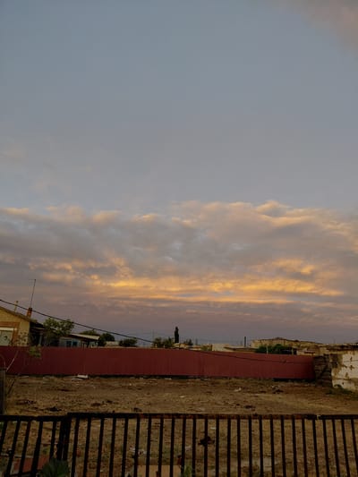 This image captures a rural landscape in Ертіс ауылдық округі, Kazakhstan, under a vast sky filled with soft, illuminated clouds tinged with orange and yellow hues, suggesting either dawn or dusk. In the foreground, a dark, metal fence with vertical bars runs horizontally across the frame, partially obscuring a patch of dry, brown earth littered with debris. Beyond the fence, a long, dark red corrugated metal fence stretches across the mid-ground. To the left, a brick building with a light-colored window is visible, adjacent to some green foliage. A tall antenna stands near the building. Several other structures, appearing to be houses or sheds with flat roofs, are scattered in the background, interspersed with trees and vegetation. The overall impression is one of a quiet, lived-in rural setting.