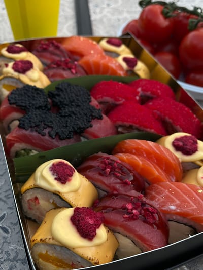 A high-angle medium shot shows a box of sushi and sashimi arranged on a patterned tablecloth. In the background, a yellow plastic cup is visible on the left, and a cluster of bright red cherry tomatoes on a vine is on the right. The sushi and sashimi are presented in a tiered arrangement within a dark gray rectangular box.
The top layer features several pieces of nigiri sushi topped with thinly sliced raw salmon. To the left of the salmon are sushi rolls with a yellow sauce and a bright red berry garnish. Behind these, there are more sushi rolls, some covered in bright red fish roe, and others topped with black caviar.
The middle layer displays more sushi rolls, some wrapped in what appears to be tuna or salmon, topped with black caviar. Next to these are several pieces of sashimi, also appearing to be salmon, arranged next to a green leafy garnish.
The bottom layer includes more sushi rolls, some with a creamy yellow topping adorned with red berry pieces. Other rolls in this layer have a glistening red topping, likely a sauce, with more red berry pieces scattered on top. There are also pieces of salmon sashimi with a reddish-pink hue, some with gold leaf accents.
The lighting suggests an outdoor or well-lit indoor setting. The overall presentation is neat and colorful, highlighting the fresh ingredients. The image does not contain any identifiable individuals or activities. The location is not specified in the image or the provided context.