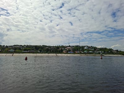 A wide shot captures a body of water with a distant, tree-lined riverbank dotted with houses and buildings. The sky is filled with a dense, patchy cloud cover, with glimpses of blue sky peeking through. Two buoys, one red and one red and white, are visible in the water. The riverbank is lined with a concrete embankment. The overall scene depicts a tranquil, rural or small-town setting.