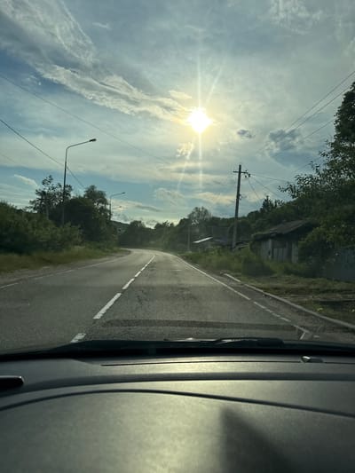 The image depicts a roadside view from within a vehicle, oriented vertically. A paved road with white dashed lines curves to the right, bordered by green vegetation and trees. In the distance, a few simple buildings are visible amongst the trees, suggesting a rural or semi-rural setting. Overhead, a bright sun shines through a partly cloudy sky, casting rays and creating lens flare. Power lines and utility poles run along the side of the road, with streetlights also visible. The scene suggests it is daytime, likely late morning or early afternoon, given the angle and intensity of the sun. The overall impression is one of a journey through a quiet, sunlit landscape. There is no visible text in the image. The location is identified as Kurinskaya, Russia.