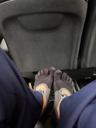 A low-angle, medium shot captures a pair of feet wearing dark gray, five-toed socks and gray slippers in what appears to be the interior of a vehicle. The socks cover the toes, with the heel and arch of the foot exposed. The slippers have the word "YOG" visible on the right foot's strap, and "GA" on the left foot's strap. The feet are positioned in front of a dark, textured surface, possibly a car's dashboard or seatback. Dark navy blue fabric, likely pants, drapes down on either side of the feet. The overall lighting is dim, suggesting an indoor or nighttime setting. No specific landmarks are visible to determine a location in Trnovo Municipality (FBiH), Bosnia and Herzegovina.