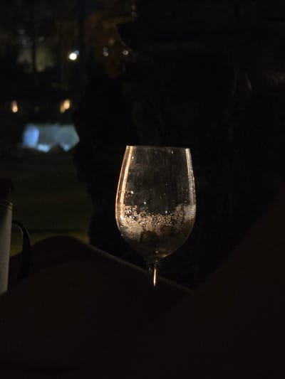A wine glass filled with a clear liquid, possibly wine or water, sits on a dark surface, possibly a lap or table, in the foreground of the image. Bubbles are visible within the liquid, clinging to the sides of the glass. The glass is illuminated from behind, creating a bright rim and highlighting the condensation on its exterior. To the left of the glass, a cylindrical object with a handle, possibly a thermos or bottle, is partially visible. The background is dark and indistinct, with faint lights suggesting an outdoor setting at night, possibly in the Hlorakas Community of Cyprus. There are no discernible people in the image.