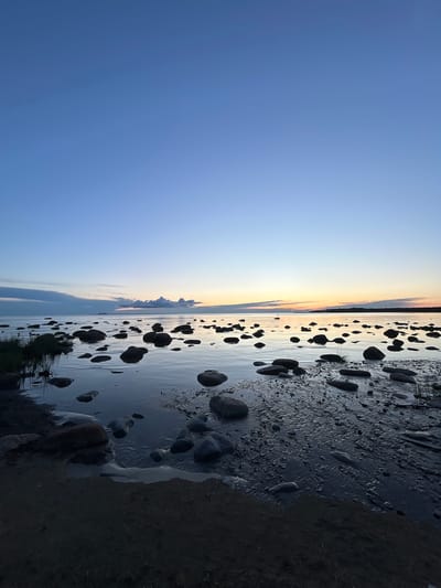A low-angle, vertical, full shot captures a rocky shore and calm sea at dusk in Haabneeme alevik, Estonia. The sky transitions from a deep blue at the top to lighter blues and oranges near the horizon. A thin band of clouds stretches across the water, with a faint, distant light source visible on the horizon, possibly a ship or building. The foreground is dominated by dark, wet sand and numerous smooth, dark stones of varying sizes. The shallow water reflects the sky and the stones. Sparse, dark green grass and reeds grow along the left edge of the frame, near the waterline. The water is still, with subtle ripples around the submerged stones. No people are present in the scene. The overall mood is serene and tranquil.