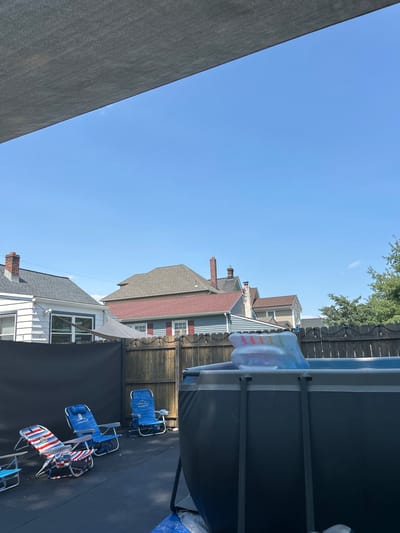 This is a photograph of a backyard in South Amboy, United States of America, on a sunny day. The scene is dominated by a large, above-ground swimming pool with a colorful inflatable toy floating inside. Surrounding the pool are several lawn chairs, including two blue chairs and one with a red, white, and blue striped pattern. A wooden fence encloses the yard, with houses visible beyond it. The sky is a clear, vibrant blue, partially obscured by a gray fabric shade structure in the foreground.