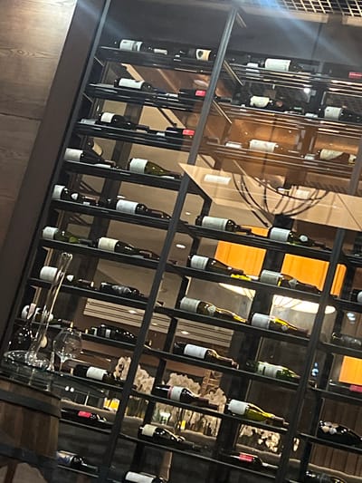 The image shows a close-up of a multi-tiered wine rack filled with numerous wine bottles. The rack is made of dark metal with shelves. The bottles are of various shapes and colors, predominantly dark glass, with some labels visible. Some bottles are partially obscured by the rack's structure. In the foreground on the left, there are wine glasses and a decanter on a shelf. The background is blurred but suggests an indoor setting with wooden elements and warm lighting, possibly a restaurant or wine cellar. The image is taken from a slightly elevated angle, looking down and to the right. No people are visible in the frame. The location is stated to be Vyshen'ky, Ukraine, but this cannot be visually confirmed from the image itself.