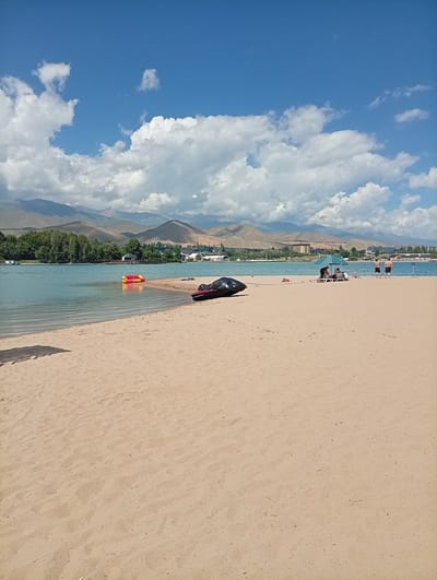 The image depicts a serene beach scene on a sunny day, likely in Çolpon-Ata, Kyrgyzstan, given the mountain backdrop. The foreground is dominated by a vast expanse of light brown sand, with some scattered debris and footprints suggesting recent activity.

In the middle ground, a calm, turquoise body of water stretches towards a tree-lined shore and distant, hazy mountains. The water is dotted with a few small boats and a brightly colored inflatable toy, consisting of red and yellow sections, floating near the shore.

Further along the beach, several individuals are visible. Under a large green umbrella, a group of people are seated on lounge chairs, relaxing. Two men are standing by the water's edge, seemingly enjoying the weather. A dark, deflated object, possibly an inflatable boat or a large bag, lies on the sand near the water.

The sky above is a vibrant blue, punctuated by fluffy white clouds that cast soft shadows on the landscape. The overall impression is one of a leisurely day at the beach, with the natural beauty of the lake and mountains providing a picturesque setting. The time of day appears to be daytime, likely late morning or early afternoon, given the bright sunlight and clear skies. No visible text is present in the image.