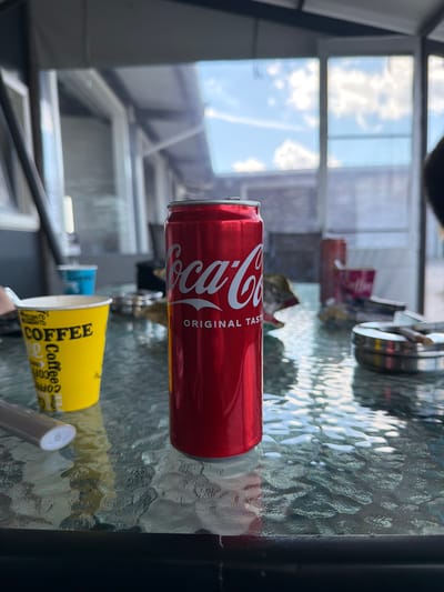 The image shows a Coca-Cola can and a yellow coffee cup on a textured glass table. In the background, there's a view through a window or enclosed patio, with structures and sky visible. The location information is unavailable, and cannot be definitively determined to be Dulovo, Bulgaria based solely on the visual content of the image. No people, subjects, or actions are visible.