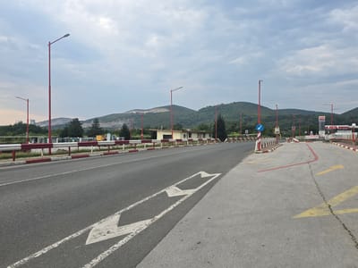 The image depicts a road scene in Zlatna Panega, Bulgaria, under a cloudy sky. A two-lane asphalt road occupies the foreground, with white directional arrows painted on its surface. The road is bordered by a red and white striped barrier and a sidewalk. In the background, a low building with a light-colored facade and a dark roof stands beside the road. Beyond the building, green, tree-covered hills rise, with some evidence of quarrying or construction on their slopes. Several red-painted lampposts are visible along the roadside. To the right, a fuel station with "AVIA" signage and a gas pump canopy is present. Traffic signs, including a blue circular sign with a white arrow and a red octagonal stop sign, are also visible. The overall scene suggests a roadside area with a commercial establishment and surrounding natural landscape.