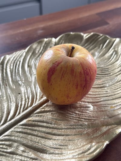 An apple rests on a golden, leaf-shaped dish. The dish is textured with raised lines that mimic the veins of a leaf. The apple is mostly yellow with streaks of red. The dish is placed on a wooden surface. The background is blurred, showing what appears to be a kitchen counter and cabinetry. The scene is indoors, likely in a home. The location in Птицевод-2 Снт, Russia is not discernible from the image.