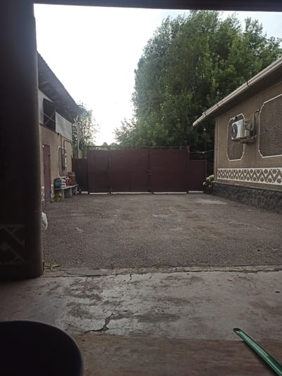 The image shows an outdoor scene in Sokuluk, Kyrgyzstan. The setting appears to be a residential area with a building on the left and a paved area on the right, possibly a driveway or yard. A large, dark brown gate is prominent, suggesting it leads to a private property or a communal area.

On the building to the left, there is an air conditioning unit mounted on the wall, which has decorative patterns along its upper edge. Next to the AC unit, a gutter runs along the roofline. Below the AC unit, there is a closed door. Further along the building, more doors and windows are visible, and some containers, possibly for storage or water, are lined up. A large tree with dense green foliage is situated behind the building, partially obscuring the sky.

The ground is covered with asphalt or a similar material, showing some cracks and wear. To the right of the paved area, there is a concrete surface with more visible cracks. A green pipe is seen at the top right of the frame, extending from the concrete. The overall impression is of a functional, perhaps slightly utilitarian, outdoor space.

The time of day appears to be daytime, with bright sky, though the lighting is somewhat muted, suggesting it might be overcast or late afternoon. No people are visible in the image, and no text is discernable. The scene conveys a sense of everyday life in a residential setting.