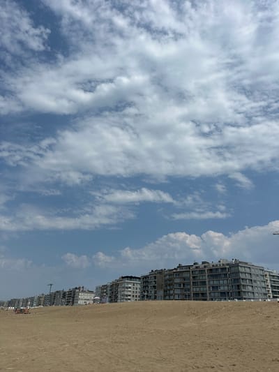 This image depicts a sandy beach with a row of tall, modern buildings lining the coast. The buildings are multi-story structures with numerous windows, suggesting apartments or hotels. The beach itself is wide and appears to be made of light-colored sand. The sky is blue with scattered white clouds, indicating fair weather, likely during the daytime. There are a few small figures of people visible on the beach, some near umbrellas and beach chairs, suggesting it is a populated area and likely a public beach. A crane is visible in the distance among the buildings, possibly indicating ongoing construction or maintenance. The orientation of the image is landscape.