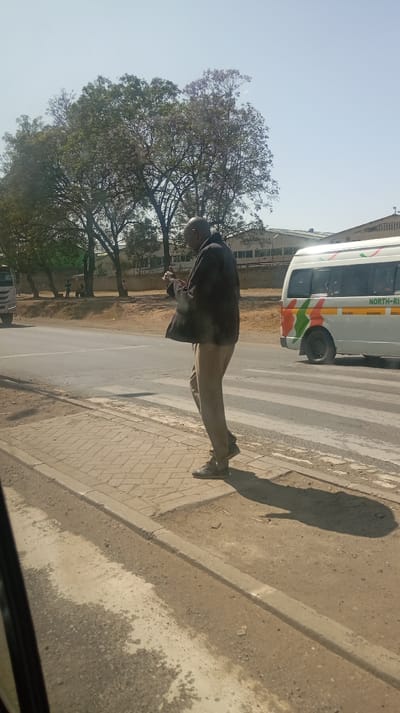 An older man in a dark jacket and light pants stands on a sidewalk, looking at a mobile phone in his hands. He is near a crosswalk on a road with vehicles, including a white van. Trees are in the background with a building in the distance. The weather appears sunny and the image is taken from inside a vehicle.