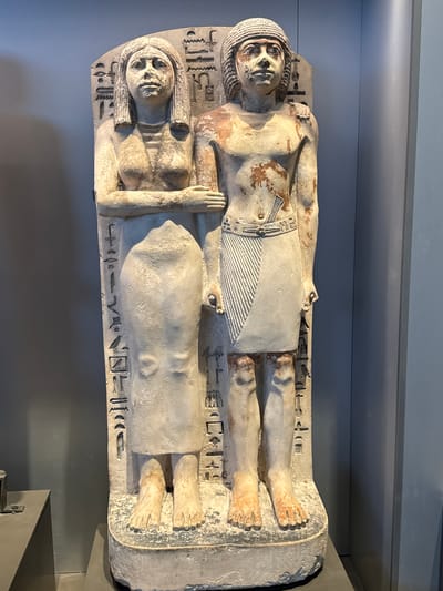 A statue of a standing man and woman is displayed against a grey background. The man is on the right, and the woman is on the left. Both figures are carved from stone and appear to be ancient Egyptian in style. Hieroglyphs are inscribed on the back of the statue. The man wears a kilt and a cap, while the woman wears a long dress. Their arms are linked. The statue is on a pedestal. The overall lighting suggests an indoor display, possibly in a museum. The image does not contain information about the city of Kafrat Nassar, Egypt.