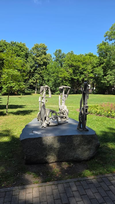 A statue composed of metal abstract figures stands on a large, rough-hewn stone base in a park. The statue depicts three figures, two seated and one standing, with elongated, angular forms. The environment is a well-maintained park with a lush green lawn, mature trees, and a clear blue sky. A paved walkway is visible in the foreground. The statue is located in Palanga, Lithuania.