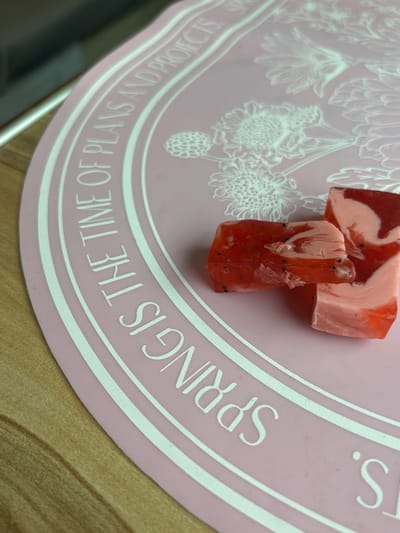 The scene presents three irregular fragments of a red, pink, and white swirled substance, consistent with confectionery or artisanal soap, positioned on a pink silicone mat. The substance displays translucent red sections containing small dark specks, intermixed with opaque light pink and white marbling. The pink silicone mat, partially visible, features white embossed text in an arc, reading "SPRING IS THE TIME OF PLANS AND PROJECTS." White embossed floral patterns, resembling chrysanthemums, are also visible on the mat's surface. The mat rests on a light-colored wooden surface, exhibiting visible grain, which occupies the lower and left portions of the frame. No individuals are present. The specific location within the city of Barabinsk, Russia, cannot be determined from the visual evidence.