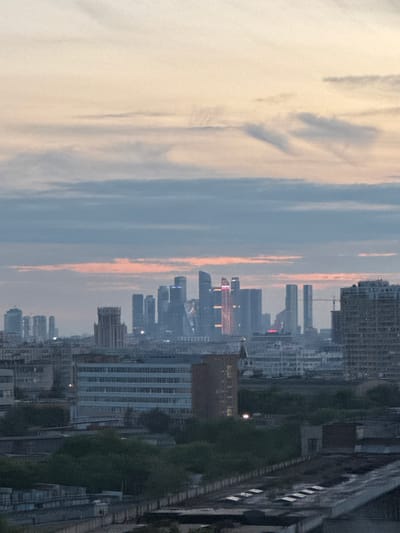 This is a landscape photograph of a city skyline at dusk. The sky is a gradient of pale yellow at the top, fading to light blue and then a band of pinkish-orange clouds, with darker blue clouds lower down. In the distance, a cluster of modern skyscrapers, some illuminated, forms the city's core. To the left and right, less prominent buildings and structures are visible, including what appears to be older architecture and industrial-looking buildings closer to the foreground. Trees and lower structures are visible in the mid-ground and foreground, suggesting a layered urban environment. There are no people or animals clearly discernible in the image. The overall impression is one of a city at the end of the day, with a sense of scale provided by the juxtaposition of the distant modern skyline and the closer, more mundane structures. The location specified as Bobrovo, Russia, does not appear to be a major city with such a prominent modern skyline, suggesting this might be an inaccurate or misattributed location.