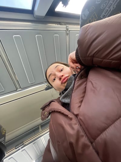The image is a low-angle selfie taken inside a public transport vehicle, likely a train or bus. A single individual, appearing to be a young adult with dark hair pulled back, is positioned in the lower-middle frame, looking directly at the camera with a slight pout. The person is wearing a grey turtleneck and a reddish-brown puffer jacket; their right hand is raised, with fingers touching the right side of their face near the ear. The vehicle's interior features light grey paneled walls with recessed vertical lines, horizontal metal railings or luggage racks, and windows showing an overcast sky. A patterned fabric seat back is partially visible in the upper right, displaying abstract illustrations of vehicles. The specific location within Fryazevo, Russia, is not ascertainable from the visual content of the image.