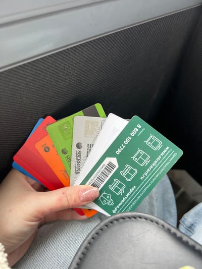 A fair-skinned hand, likely female, with short, manicured nails painted with glittery pink polish, holds a fan of approximately eight plastic cards in the central foreground. Minor red bumps are visible on the thumb of the hand. The cards display various colors including blue, red, orange, green, grey, and white. Identifiable cards include a grey Sberbank card and a prominent green "Strelka" Unified Transport Card for Students of the Moscow Region. This green card features a barcode, the phone number 8 800 100 7790, the website www.strelkacard.ru, and icons representing various modes of transport. The individual's leg, clad in light blue denim jeans, is partially visible beneath the hand. A portion of a dark, stitched bag or purse is present in the lower right. The background consists of a dark, textured surface with horizontal ribbing, consistent with an interior panel, topped by a smooth, lighter grey stripe, suggesting an indoor environment, possibly within a vehicle. The image does not provide information to confirm the specific location within Lipitsy, Russia; rather, one card explicitly references the "Moscow Region."