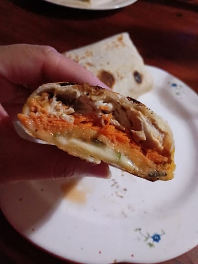 A person is holding a half-eaten burrito in their right hand. The burrito is filled with chicken, carrots, and a white sauce. It is resting on a white plate with a floral pattern. The plate is on a wooden table. The burrito appears to be in the process of being eaten. The coordinates provided (latitude: -34.473, longitude: -58.639) correspond to a location in El Talar, Argentina.