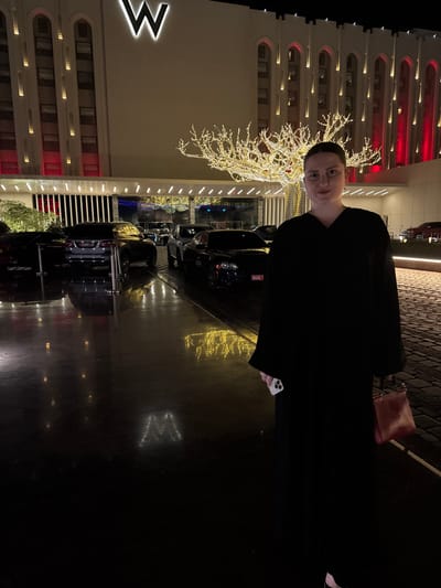 A person in a black robe is standing in front of a building at night. The building has multiple floors with illuminated windows and a large "W" logo. Decorated trees with lights are on the property. Several dark-colored cars are parked in front of the building. The ground is reflective, mirroring the building's lights and design. The person is holding a white phone in their right hand.