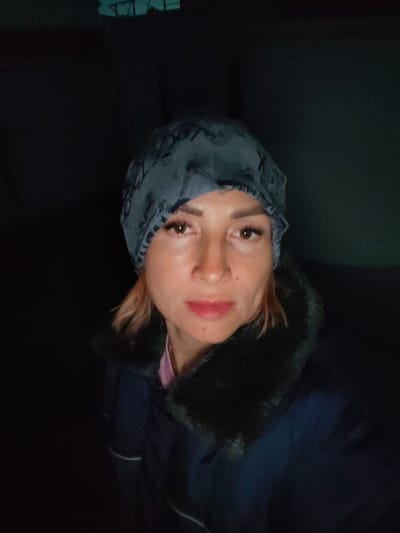 A medium close-up selfie shows a woman with light skin and reddish-blonde hair, wearing a grey patterned beanie and a dark blue jacket with a dark fur collar. Her head is tilted slightly to her left, and she is looking directly at the camera with a neutral expression. Her makeup includes mascara and lip gloss. The background is dark and indistinct, with a faint hint of a dark green wall on the right side, and a light structure with some geometric shapes visible at the top. There is no discernible indication of the location being Irdomatka, Russia.