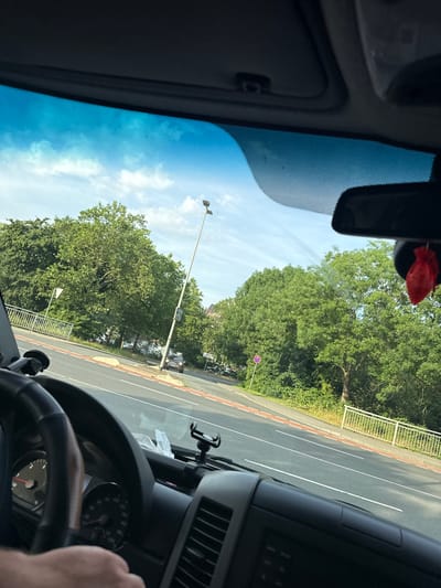 The image is a first-person view from inside a vehicle in Wetzlar, Germany. The driver's hands are visible on the steering wheel, and the dashboard with instruments is in the foreground. Outside the windshield, a road is visible with lane markings and a sidewalk. Lush green trees line the side of the road, and a street light is present. In the background, there are buildings and a clear sky with some clouds. A red car air freshener is hanging from the rearview mirror.