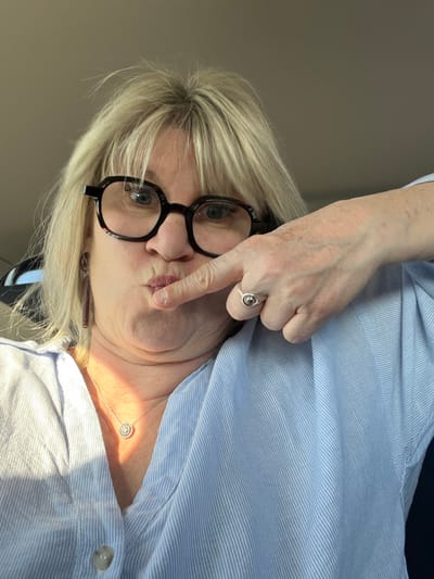 A female individual is depicted in a close-up, chest-up view, appearing to be inside a vehicle in Étoile-sur-Rhône, France. The subject has fair skin, light blonde hair with bangs, and wears dark-framed, circular spectacles. Her lips are pursed forward in a pouting expression, with her right index finger positioned against the center of her lower lip. She wears a light blue, vertically striped collared shirt. Visible accessories include a dark, elongated dangle earring on her left ear, a silver-toned chain necklace with a circular, jeweled pendant, and a silver-toned ring with a spherical central element on the ring finger of her right hand. The background is a uniform, plain light brown or beige color, consistent with an interior ceiling or headliner of a vehicle. Light is cast from the left, creating a shadow across the lower-right portion of her face and shirt.