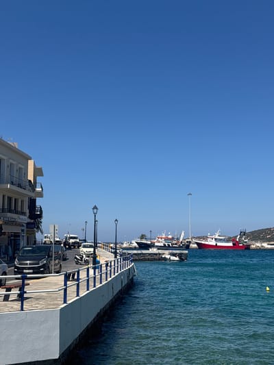 This image depicts a scenic view of a harbor in Agios Nikolaos, Greece, under a clear blue sky, suggesting a sunny day. The perspective is from a quay, looking out towards the water and a line of docked boats and ships.

Dominating the foreground is a paved quay with a white and blue railing. Several vehicles are parked along the quay, including a black SUV, a white van, and other cars. The quay appears to be a bustling area with lampposts lining its edge.

Further out in the water, several boats and ships are moored. A large red and white vessel is prominently visible, alongside smaller fishing boats and leisure craft. The water is a clear, rippling blue-green, indicating calm conditions. In the distance, the coastline is visible, with hills and buildings suggesting the presence of a town or city. A yellow buoy floats in the water.

The scene suggests a typical day at a Greek harbor, with vessels ready for departure or return. The clear sky and bright sunlight point to the time of day being either morning or afternoon. The overall atmosphere is one of a vibrant, active port. Visible text on a sign includes "NOSTOS CRUISES" and "SPINALONGA BOAT TRIPS & TICKETS," further confirming the location and suggesting tourist activities.