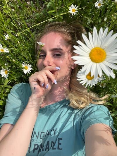 A young woman with blonde hair is lying on her back in a grassy field dotted with daisies. She is wearing a teal t-shirt with black lettering that reads "SUNDAY MAX & SURF PRAIZE". Her right hand is raised to her face, with her fingers touching her lips. The woman's nails are painted a light lavender color. She has a small tattoo on the back of her left hand. The sun is casting shadows across her face and body, indicating it is daytime. The background is filled with green grass and more daisies. The location is identified as Yasnoe, Russia.