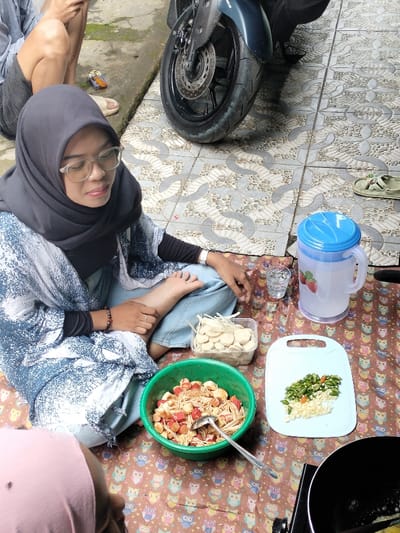 The image depicts a casual outdoor or semi-outdoor scene in Leuwiliang, Indonesia, centered around food preparation. A woman, wearing a black hijab, clear-rimmed glasses, a blue and white patterned top over a black long-sleeved shirt, and light blue pants, is seated cross-legged on a colorful mat adorned with an owl pattern. She appears calm and content, wearing a wristwatch, a ring, and a bracelet.
Surrounding her are cooking ingredients: a green bowl holds a mix of red sausage-like pieces, white meatballs/mushrooms, and thin white strands, with a ladle in it. A clear plastic container nearby contains sliced white items, likely meatballs or mushrooms. On a white cutting board, there's a pile of chopped green chilies and minced garlic or onions. A small clear glass of water and a larger clear pitcher with a blue lid and red strawberry decals are also present on the mat. Part of a black cooking pot or pan is visible in the bottom right, suggesting active cooking.
Other individuals are partially visible: legs and a hand of a person in shorts are seen to the upper left, near a cigarette pack, and another person in a pink top is in the bottom left. The ground features patterned grey and white floor tiles. The front wheel and fender of a dark-colored motorcycle, with the brand "YAMAHA" faintly visible, are in the background. A pair of light-colored sandals rests on the tiled ground. The scene appears to take place during the daytime under bright, ambient light, suggesting a warm and pleasant day for this communal cooking activity.