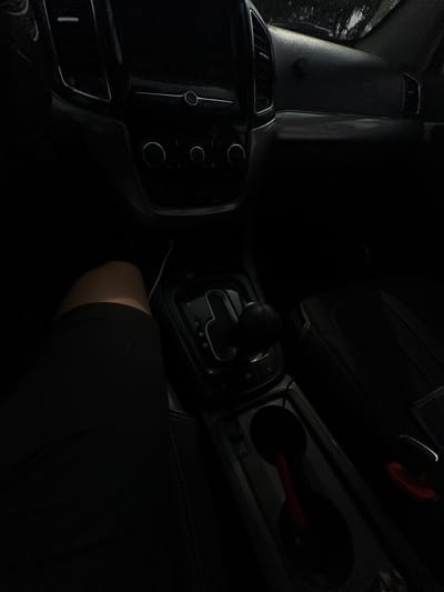 The image shows the interior of a car, likely taken from the perspective of a passenger. The scene is dimly lit, suggesting it might be nighttime or a heavily shaded interior. Dominating the foreground is a person's leg, covered in black fabric, suggesting they are wearing trousers or leggings. The central focus is the car's gear shift, which is in the "N" (Neutral) position. Adjacent to the gear shift are what appear to be climate control buttons and a part of the infotainment system, also mostly in shadow. To the right, a cup holder is visible, with red accents that hint at the car's interior design. The overall impression is one of a still moment within a vehicle, with the low light and focus on the controls adding a sense of quiet or perhaps anticipation. The mention of Istiqlol, Uzbekistan, provides geographical context, although there are no overt visual cues within the image to confirm this specific location, beyond it being a car interior.
