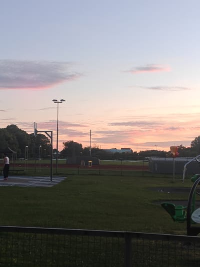 The image captures a sunset view over a public park in Worthing, United Kingdom. The sky is painted with hues of pink and orange, with scattered clouds. In the foreground, a black metal fence separates the viewer from a grassy field. To the left, a basketball hoop and court are visible, with a person in a white top and black trousers standing on the court. Further back, there are trees, sports facilities including a running track, and some buildings. The overall scene depicts a serene evening in an outdoor recreational space.