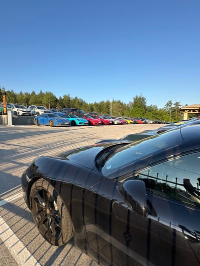 A long line of brightly colored sports cars, predominantly Porsches, are parked in a lot with a forest in the background and a clear blue sky above. The foreground is dominated by the front section of a black Porsche, its wheel and side mirror visible. The other cars, in shades of blue, teal, red, yellow, and white, are lined up in the middle ground, some parked on a paved surface and others on what appears to be a raised area. The scene suggests a car meet or a gathering of enthusiasts. The location is specified as Kerenite, Bulgaria.