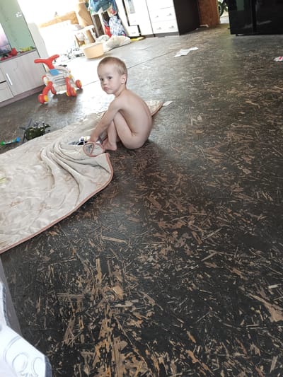 A young, light-skinned child, possibly a toddler, sits on a dark, patterned floor in what appears to be an indoor setting in Teykovo, Russia. The child is wearing only a diaper and is looking towards the viewer with a neutral expression. To the child's left, partially covered by a light-colored blanket or quilt, are some toys including a green toy helicopter and a white toy police car. The blanket also extends to the foreground on the left.

In the background, a cluttered room is visible. There's a white cabinet or dresser to the left with various items on top, including a red object. A red and white toy with wheels (possibly a ride-on toy or walker) is near the cabinet. Further back, there are more shelves or storage units with clothes and other household items. To the right, part of a dark-colored appliance (possibly a refrigerator) is visible. The floor has a textured appearance, resembling pressed wood or particle board with dark and lighter brown tones. The lighting suggests it is daytime, likely indoors. No visible text is clearly legible in the image.